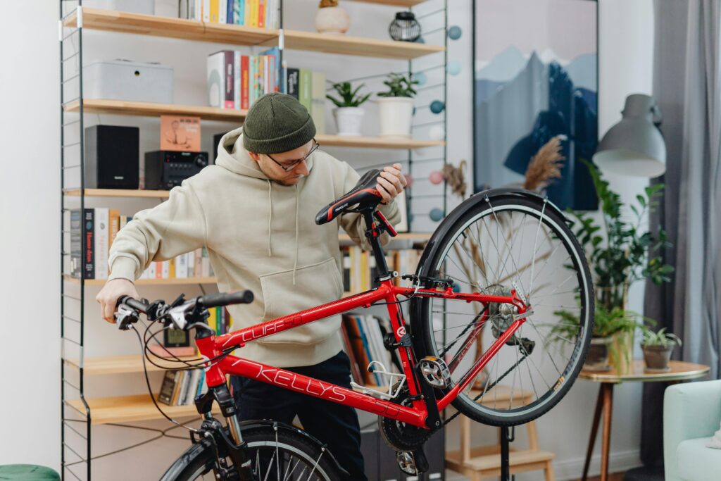Un homme en sweat à capuche répare un vélo rouge dans un intérieur chaleureux, agrémenté d'étagères et de plantes.